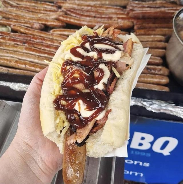 Loaded sausage in a roll with bacon, cheese and sauces, fresh off the hotplate