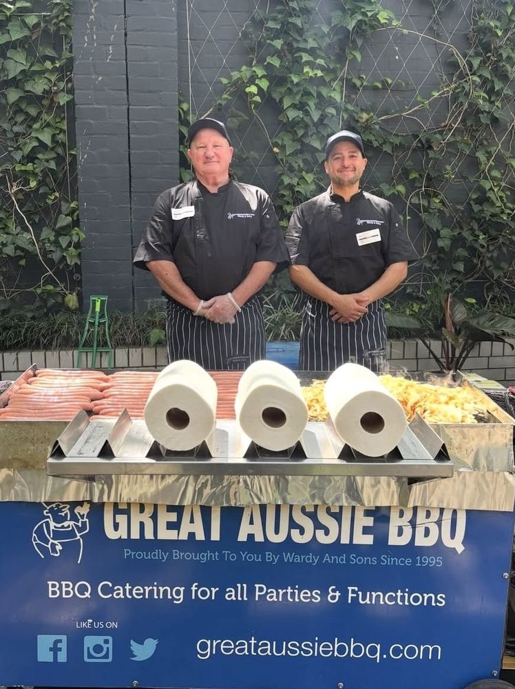 Great Aussie BBQ team members in black chef jackets behind the Great Aussie BBQ cart, its hotplate loaded with sausages and onions